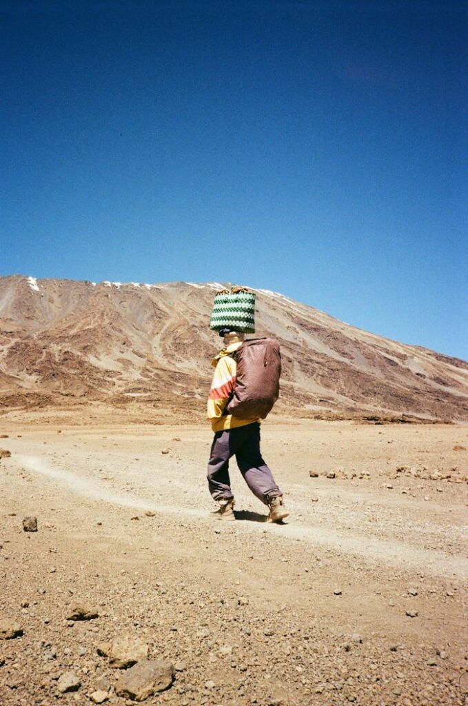 A young porter carries goods on a trek up Mount Kilimanjaro under a clear blue sky.
